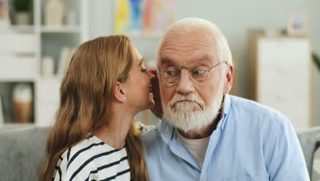 Close-up-of-the-young-blond-teen-girl-whispering-to-the-grandfather's-ear-in-the-cozy-living-room.